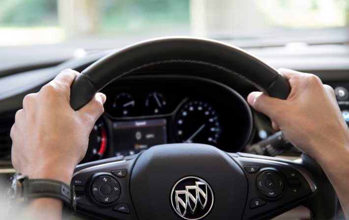 Close-up of a person's hands gripping the steering wheel of a Buick vehicle, with the dashboard and instrument panel visible in the background.