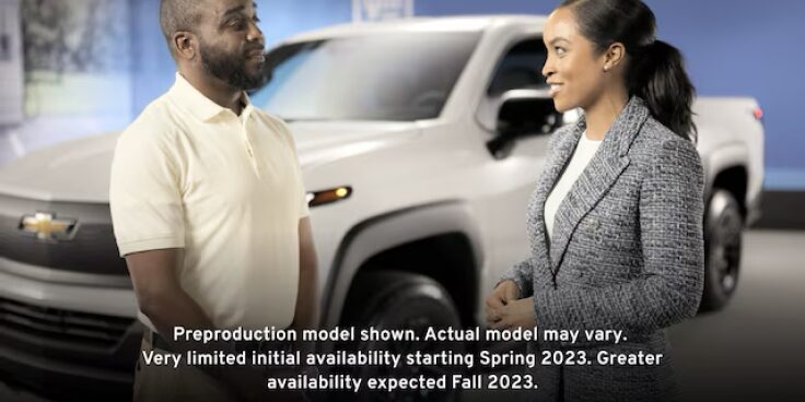 A man and woman stand next to the exterior  of the Summit White 2024  Silverado EV discussing its capabilities.