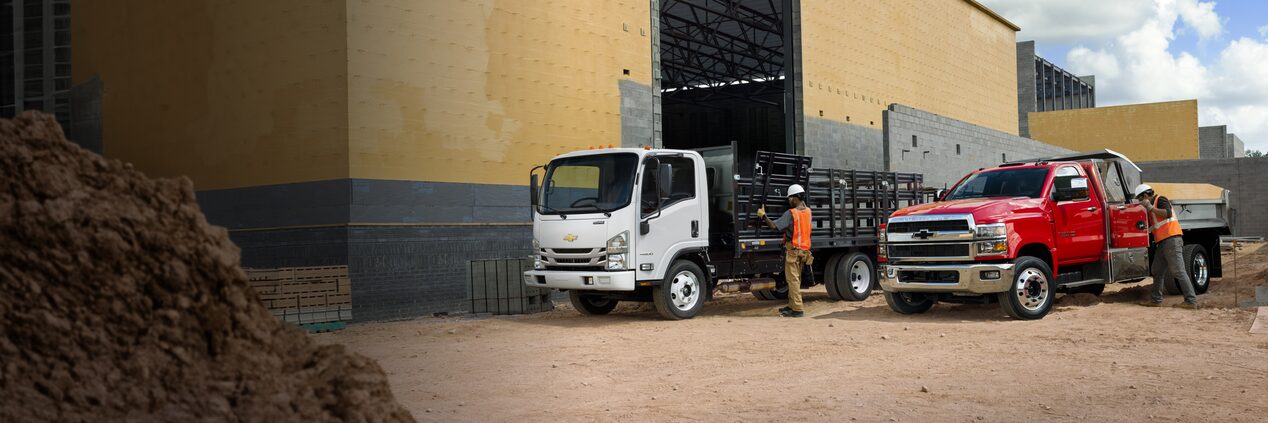 A 2023 Chevrolet Silverado 3500 HD Chassis Cab/GMC Sierra 3500 Heavy Duty Chassis Cab and a 2023 Chevrolet Low Cab Forward 6500 XD/7500 XD Truck With Platform Upfit Application on a job site.