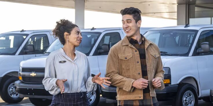 A car dealer talking to a potential customer in the lot infront of three white Chevrolet vehicles.