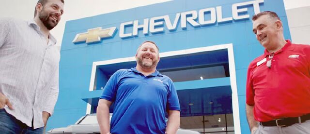 Three men standing in front of a Chevrolet dealership