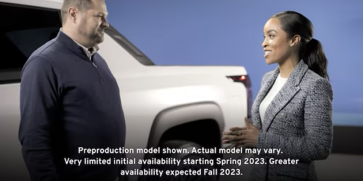 A man and woman stand next to the exterior  of the Summit White 2024  Silverado EV discussing its capabilities.